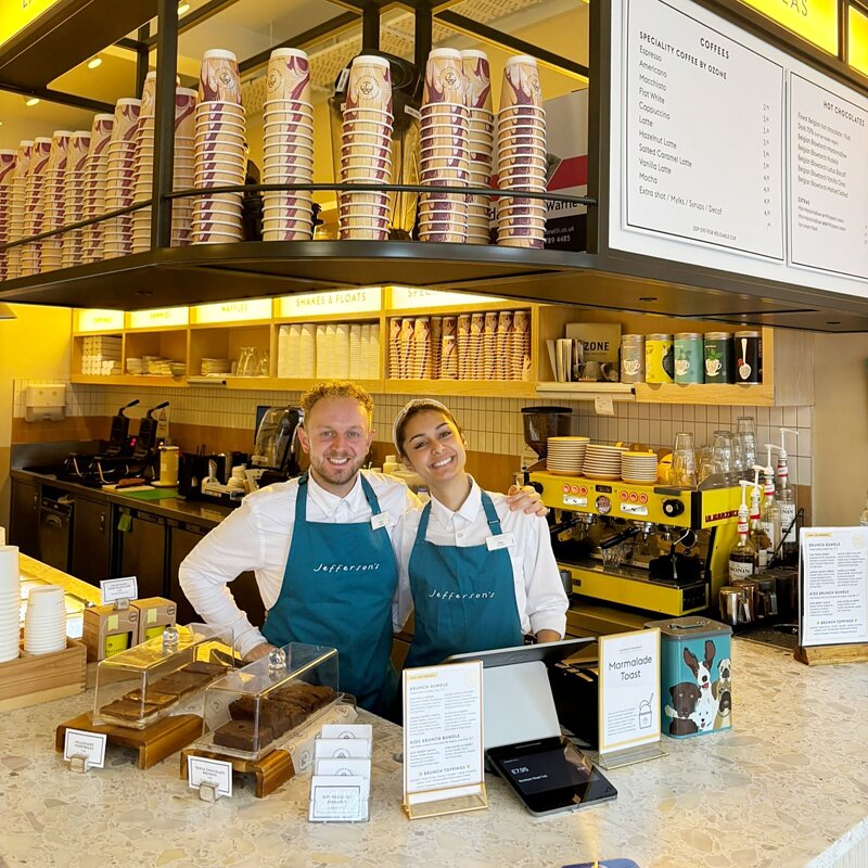 Two smiling staff in teal aprons behind the counter of the ice cream parlour, with stacked cups and coffee machines around them