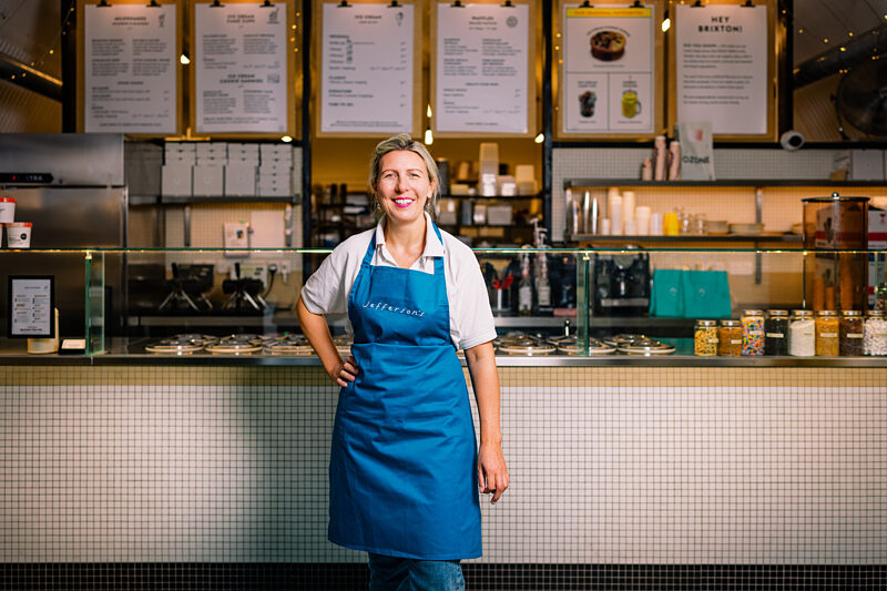 A smiling woman in a blue Jefferson's apron standing in front of the ice cream counter and menu boards at Jefferson's Ice Cream in Brixton