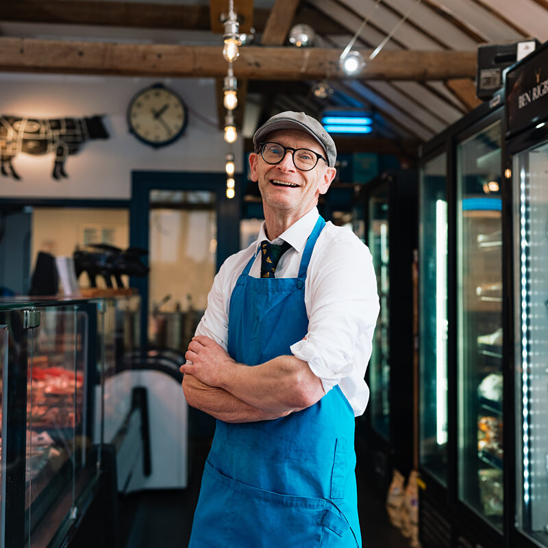 A smiling butcher in a flat cap, glasses and blue apron standing with arms folded between the meat counter and the fridges at Morgan's Butchery